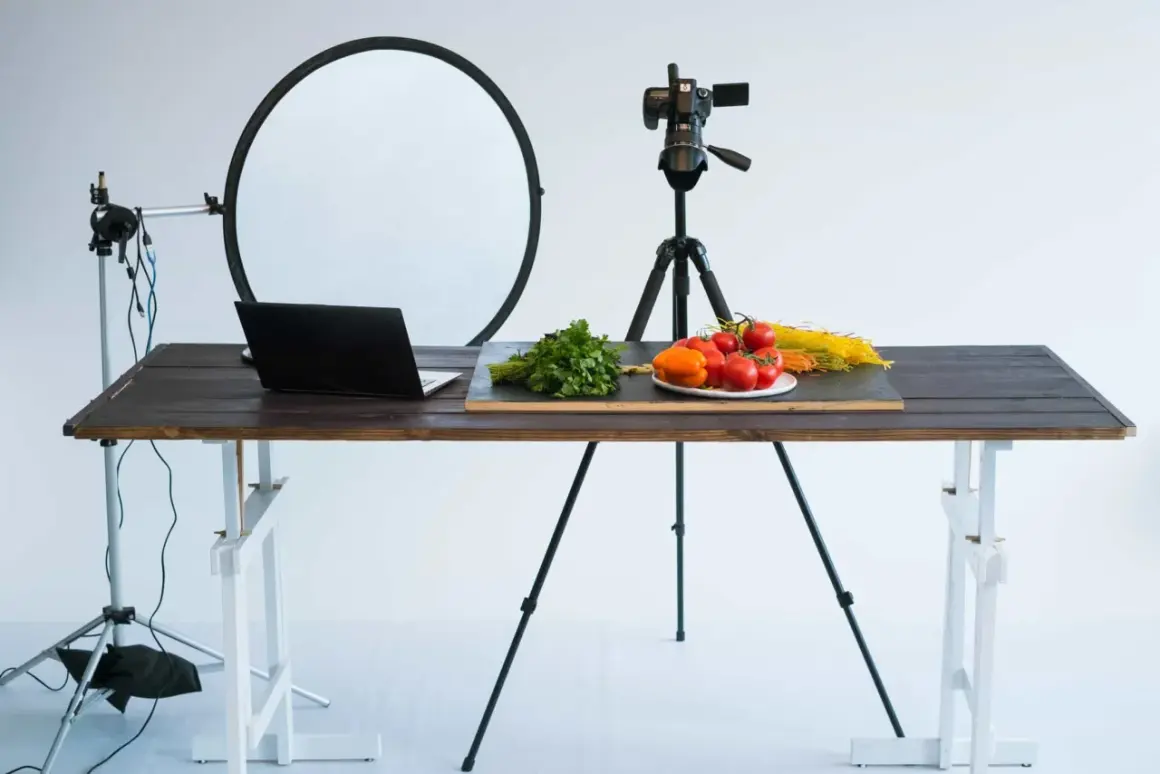 A tabletop photography setup with a camera on a tripod, a laptop, and fresh ingredients like tomatoes and pasta on a wooden table.