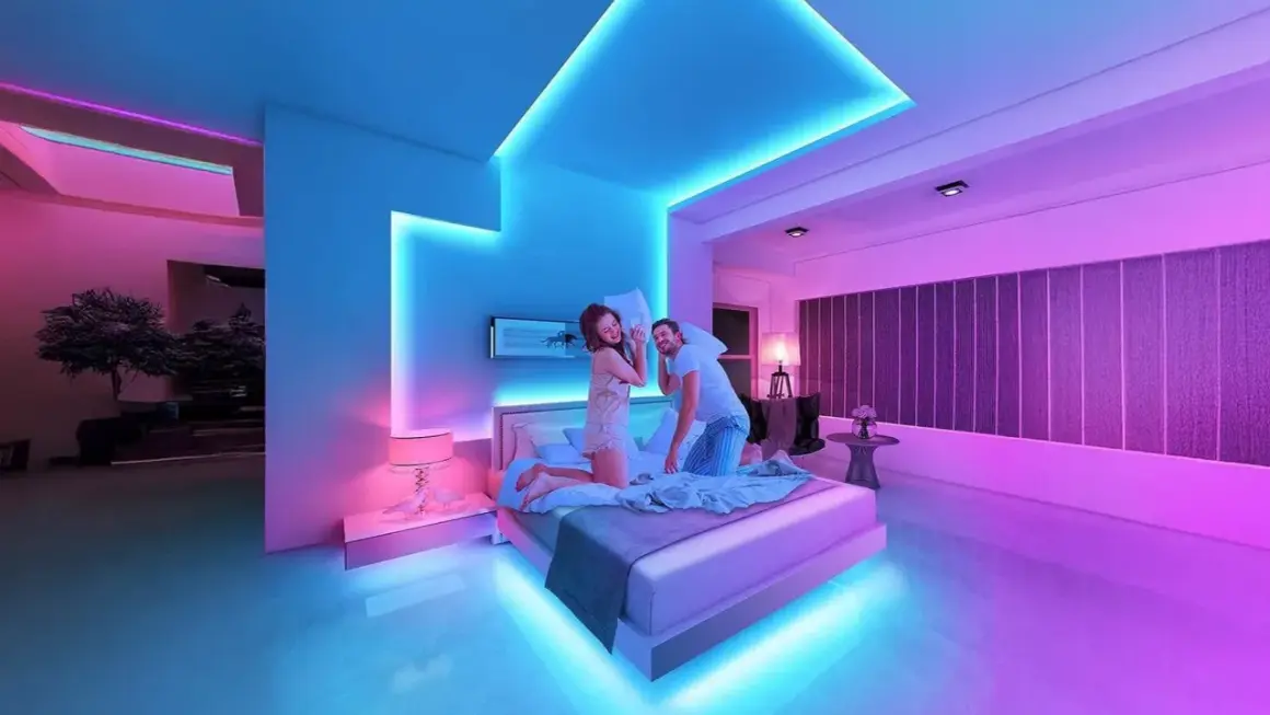 A couple has a pillow fight in a bedroom bathed in vibrant RGB light ideas, with blue and pink hues illuminating the modern space.