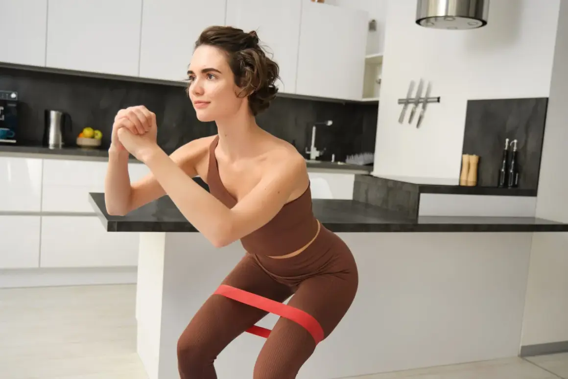 Femme en tenue de sport marron effectuant un squat avec un élastique rouge autour des cuisses dans une cuisine moderne.