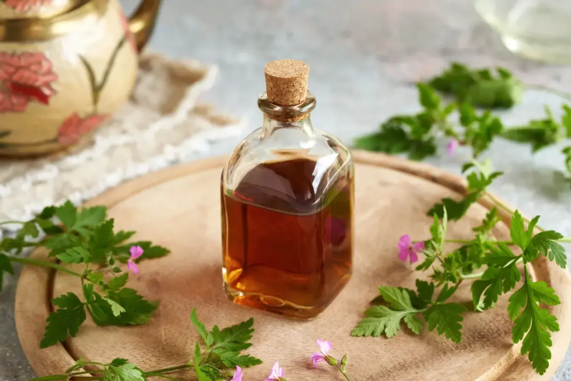 Szklana butelka z bursztynową nalewką z geranium, otoczona świeżymi gałązkami i kwiatami tej rośliny, na drewnianej desce.