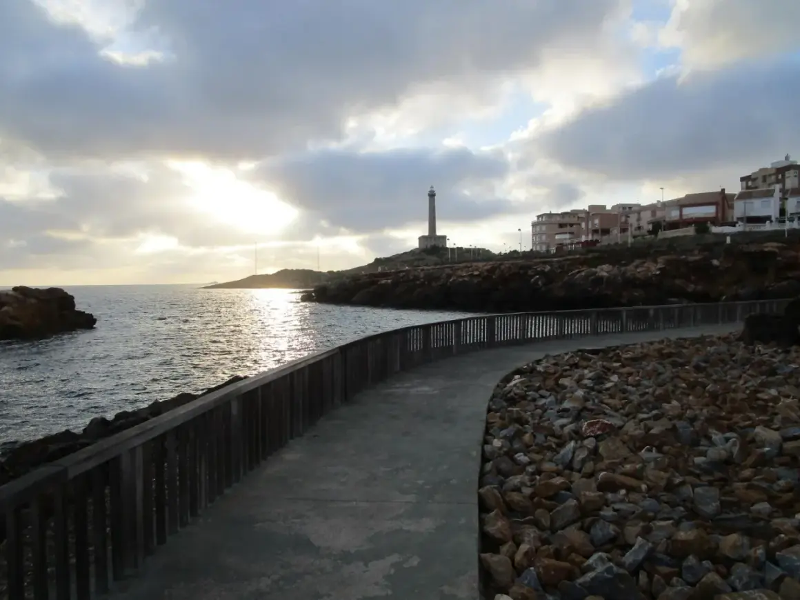 Camino de piedra y madera bordeando el mar, con un faro al fondo. La carretera subida al faro invita a pasear al atardecer.