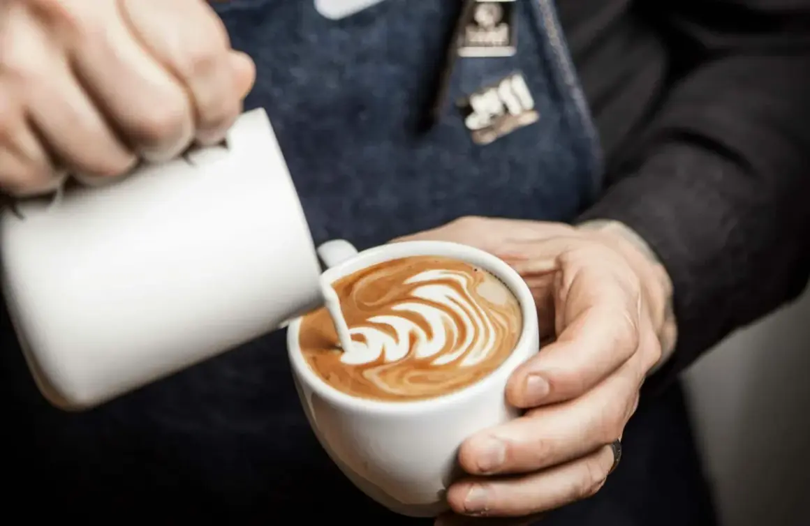 Barista skapar vacker latte art i en vit kopp. Mjölkskummet bildar ett mönster av virvlar.