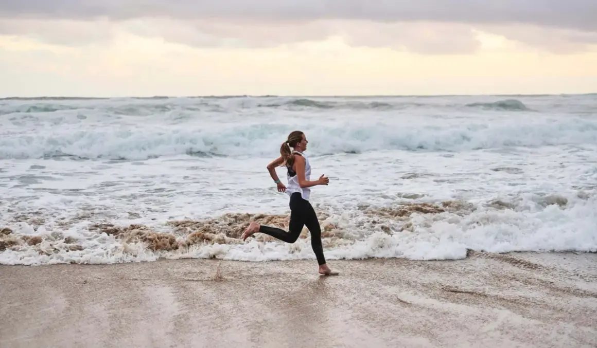 Frau joggt barfuß am Strand entlang, während die Wellen des Meeres an den Sand spülen. Sie genießt ihr tägliches Joggen.