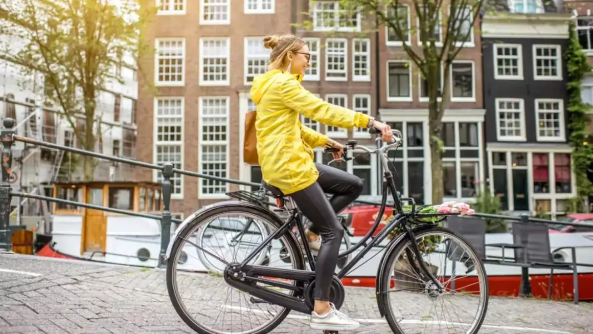 Vrouw in gele jas rijdt op een simpele stadsfiets door Amsterdam, met bloemen in het mandje.