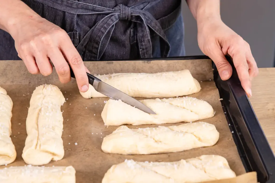 Hände bereiten Teiglinge für Laugenstangen zum Aufbacken vor. Ein Messer schneidet in einen Teigling, grobes Salz liegt bereit.