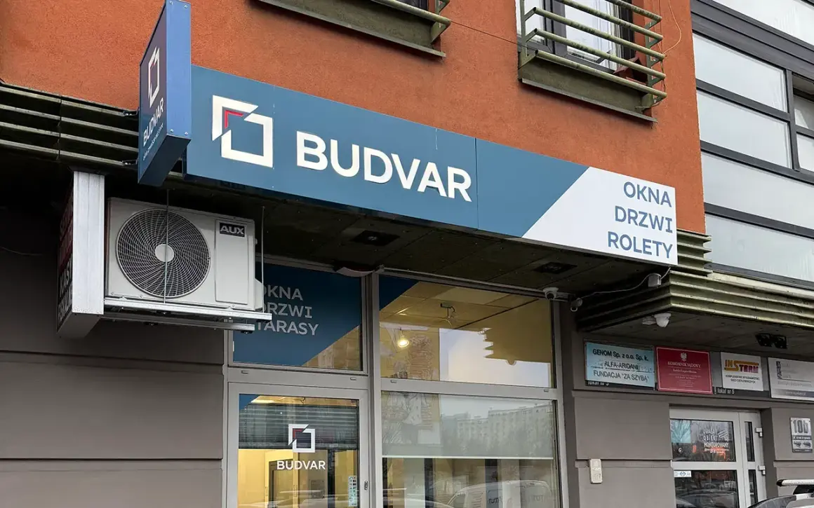Logo firmy BUDVAR z napisem "OKNA DRZWI ROLETY" na tle pomarańczowej ściany.