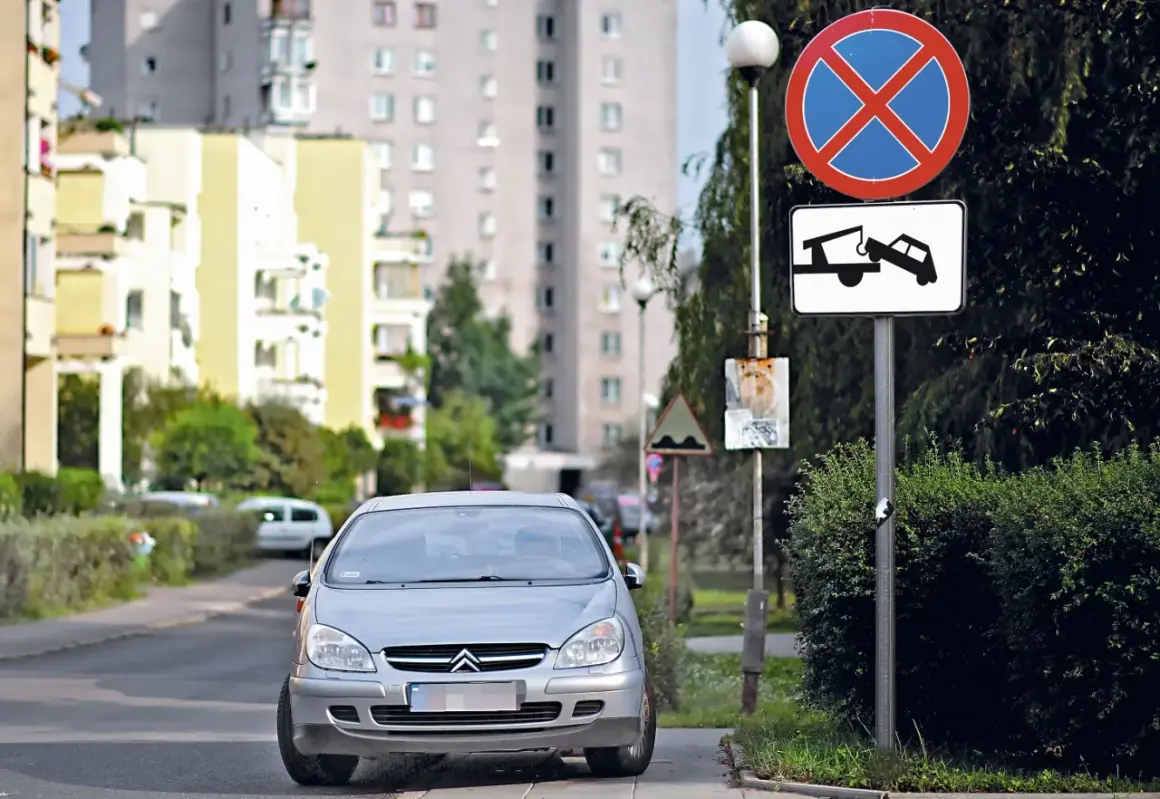 Srebrny samochód na drodze wewnętrznej, obok znaki zakazu zatrzymywania się i holowania. W tle bloki mieszkalne.