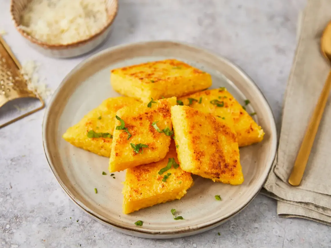 Gebratene Polenta-Ecken, italienisch garniert mit Petersilie, auf einem Teller. Daneben eine Reibe mit Parmesan.
