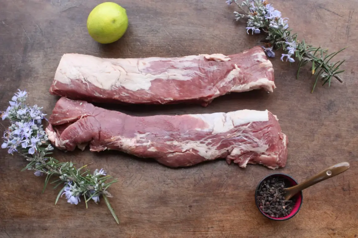 Deux pièces de filet mignon cru sur une planche en bois, accompagnées de romarin fleuri, d'une lime et de baies poivrées.