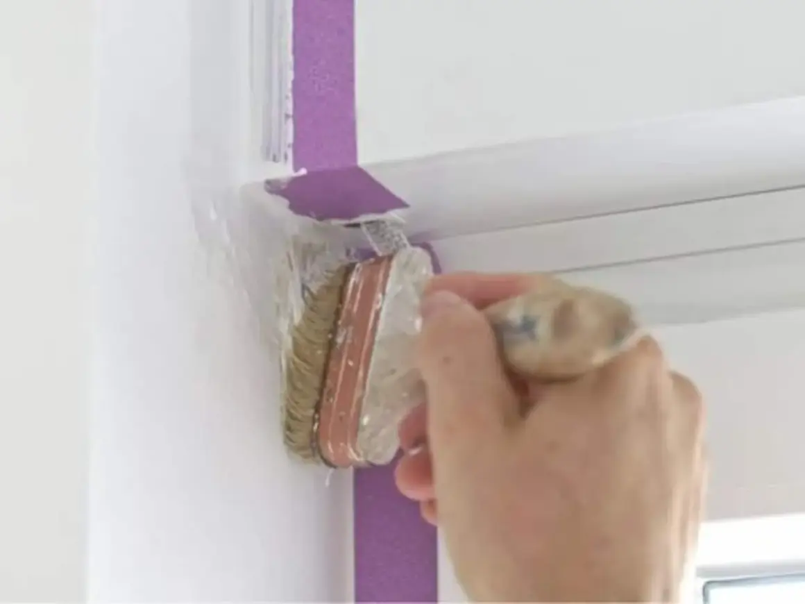 Hand streicht mit Pinsel und Klebeband eine weiße Wand.