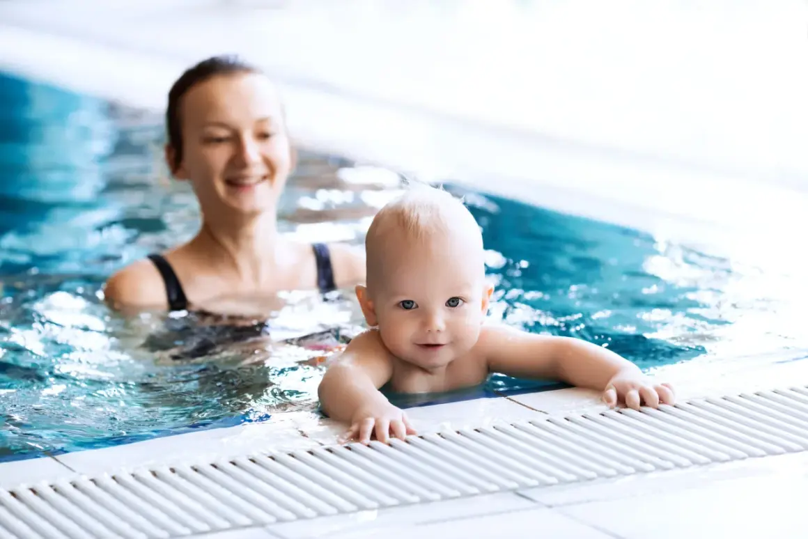Uśmiechnięty niemowlak w basenie, trzymający się brzegu. Mama w tle obserwuje go z radością.