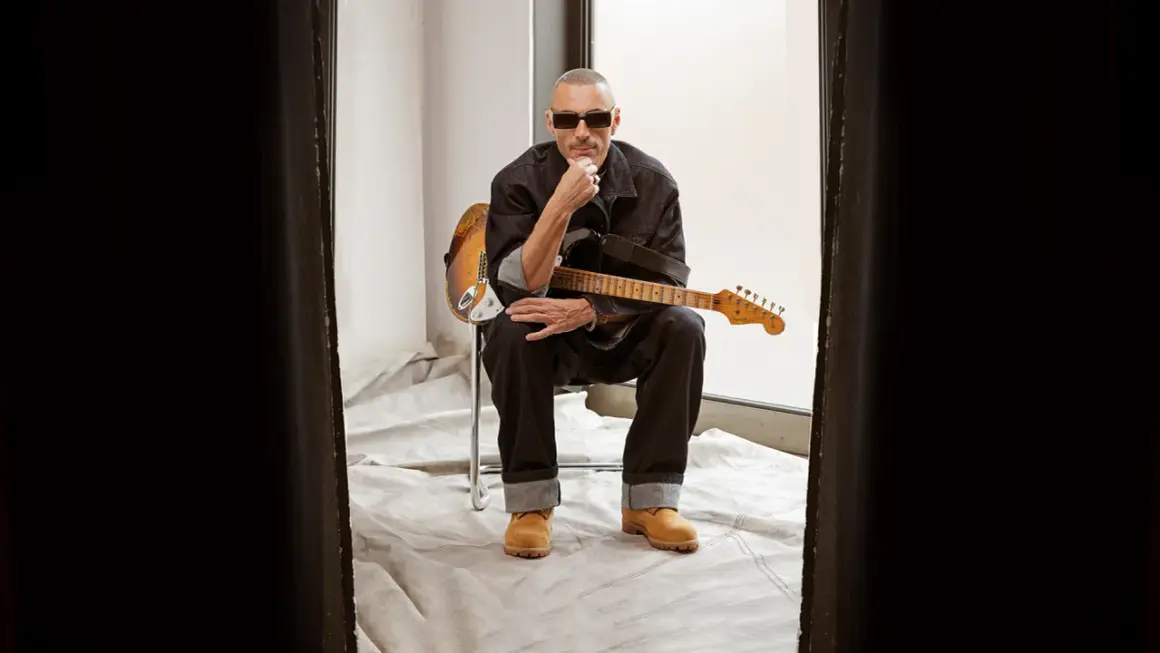 Alex Britti, con occhiali da sole e chitarra, sembra pensieroso, quasi gelido, mentre riflette sui suoi testi.