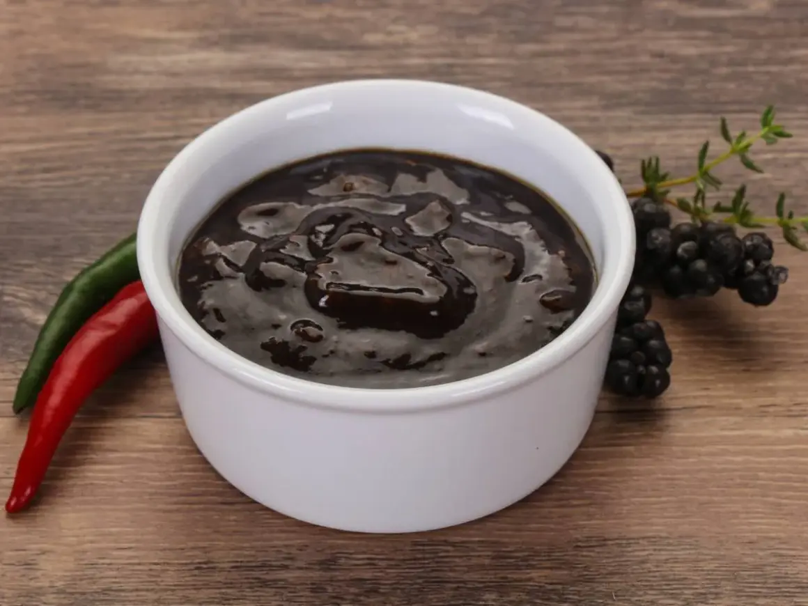 Gęsty, ciemny sos pieprzowy w białej miseczce, obok czerwona i zielona papryczka chili oraz gałązka jeżyn.