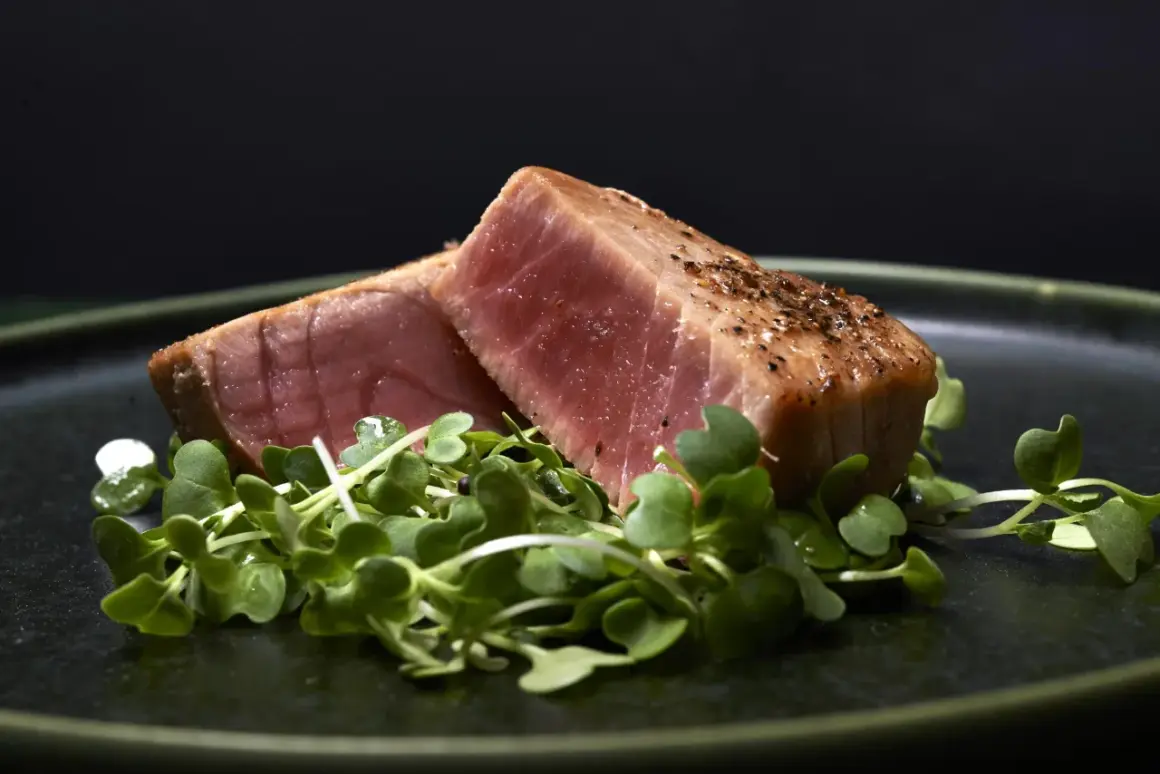 Två bitar grillad tonfisk med peppar ligger på en bädd av gröna skott på en mörk tallrik.