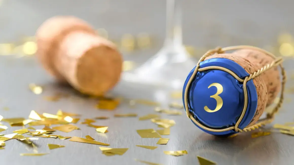 Sektkorken mit blauer Kappe und goldener "3" symbolisiert die Bedeutung des 3. Hochzeitstags. Goldkonfetti und ein weiterer Korken im Hintergrund deuten auf eine Feier hin.