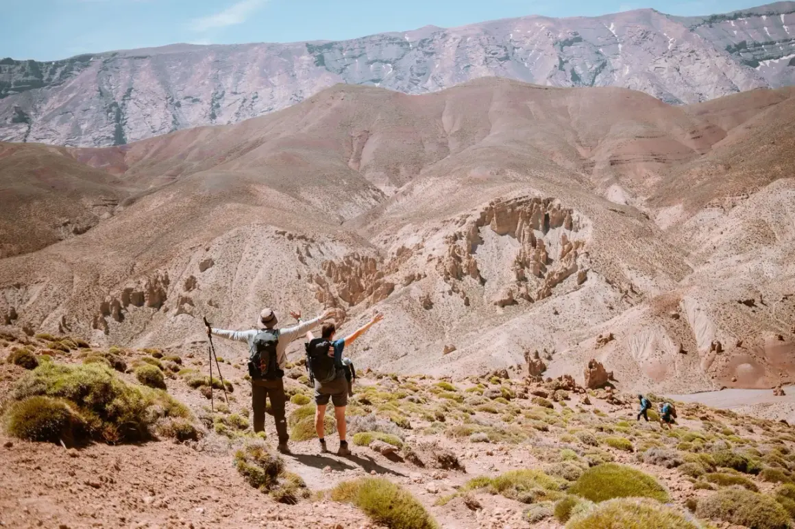 Deux randonneurs bras ouverts contemplent un paysage désertique spectaculaire lors d'une randonnée Atlas Maroc. D'autres marcheurs sont visibles au loin.