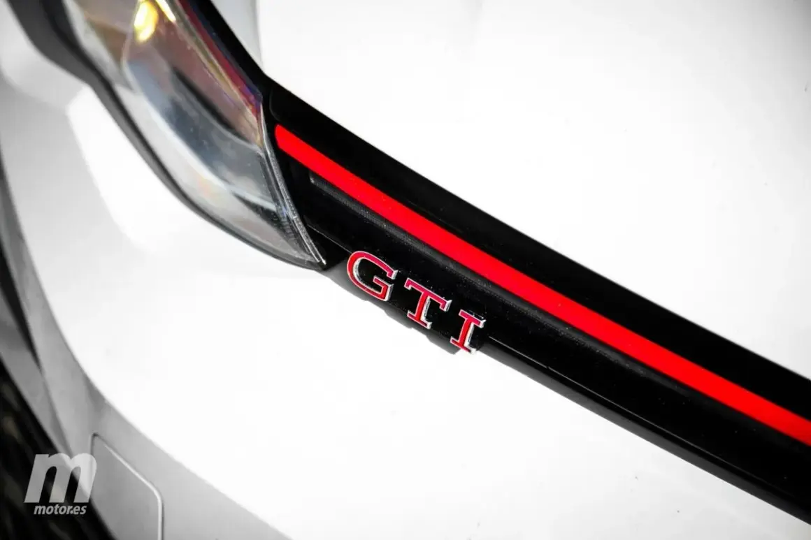 Detalle de un coche blanco con las siglas GTI en rojo y cromo, sobre una franja negra y roja.