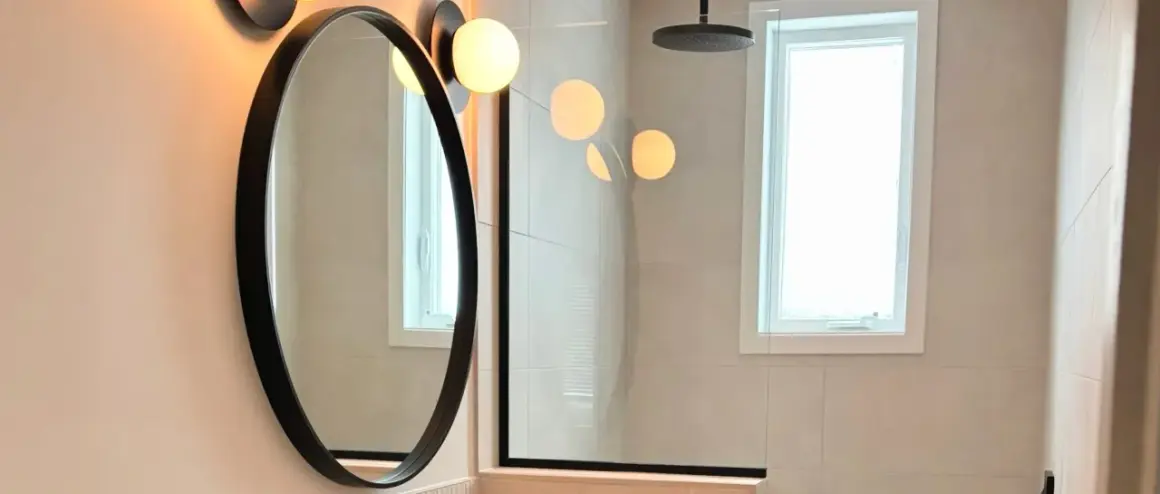 Un élégant miroir au-dessus du lavabo reflète des lumières douces. La salle de bain moderne comprend une douche vitrée et une fenêtre lumineuse.