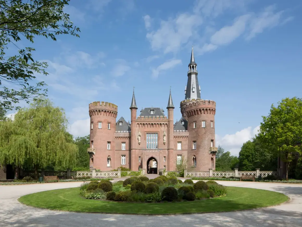 Schloss Moyland, ein imposantes Backsteinschloss mit Türmen und einem hohen Spitzdach, thront über einer gepflegten Gartenanlage mit einem runden Blumenbeet.