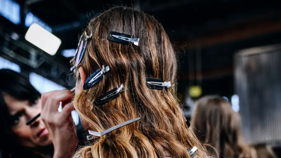 Detrás de cámaras, una maquilladora aplica producto para alisar el pelo de una modelo, sujetado con pinzas.
