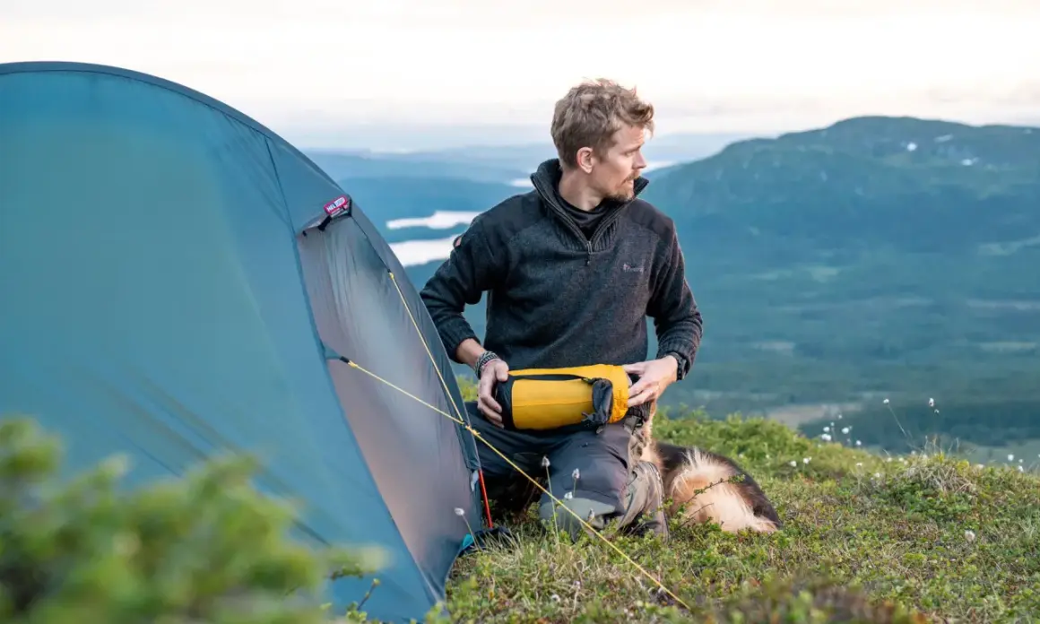 Man med hund förbereder tältet för en vandring i Norrland. Vacker fjällvy i bakgrunden.