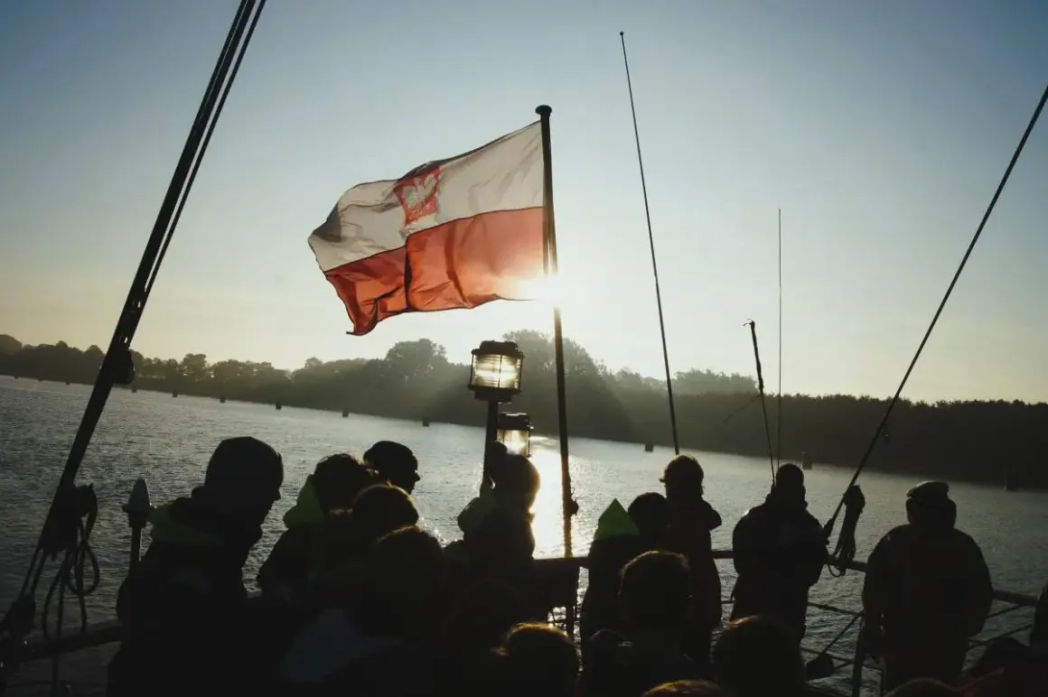 Słońce przebija się przez biało-czerwoną bandera polską na statku. Grupa ludzi obserwuje zachód słońca nad wodą.