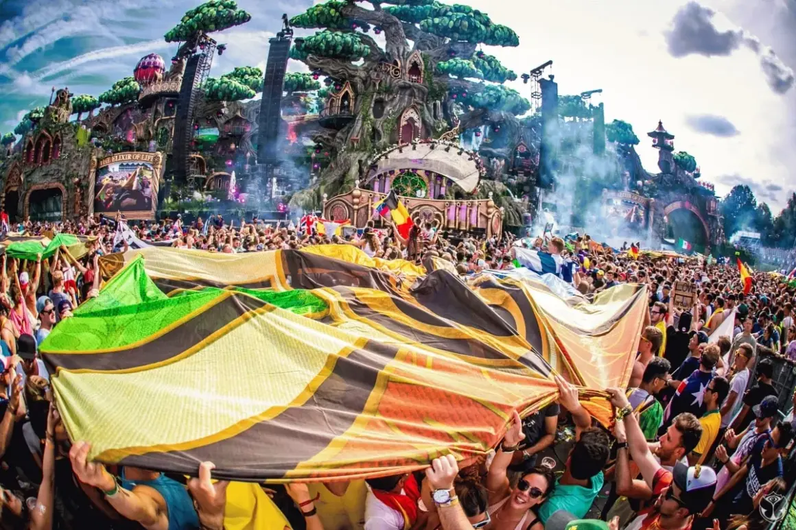 Tłum ludzi na festiwalu Tomorrowland, trzymający kolorową flagę, z imponującą sceną w tle.