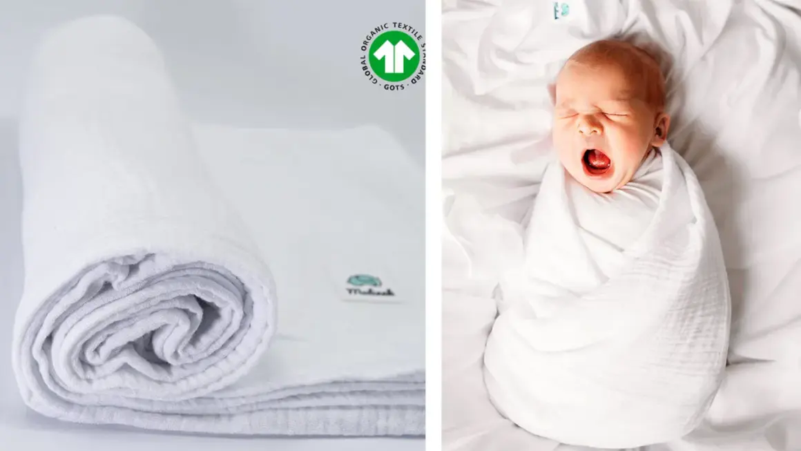 Biały, zwinięty otulacz muślinowy obok śpiącego niemowlaka. Certyfikat GOTS potwierdza jego ekologiczne pochodzenie.