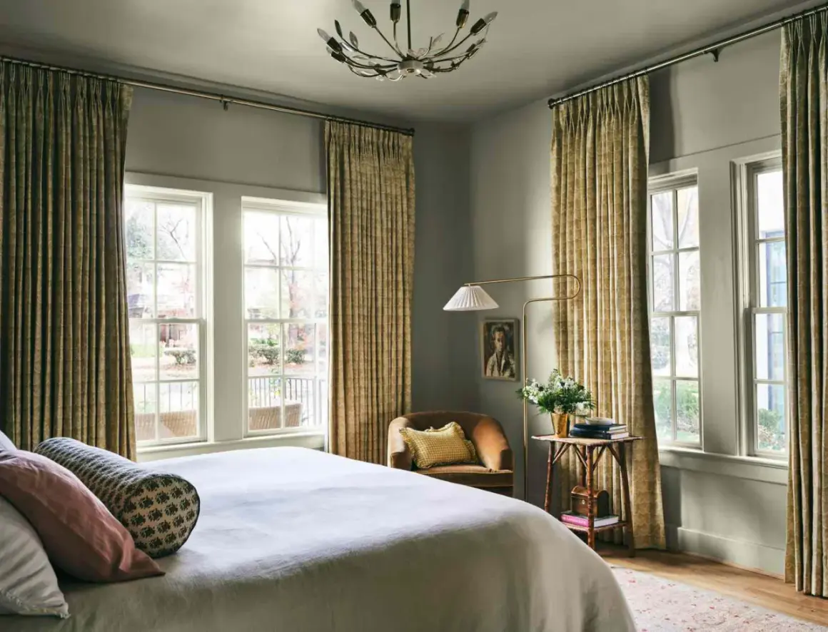 A cozy bedroom with a bed, a chair, and a floor lamp. The curtains are a beautiful millenial green, adding a touch of nature to the room.