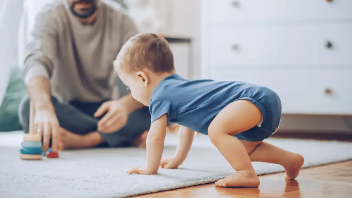 Dziecko w niebieskim body raczkuje po dywanie, obok siedzi ojciec z zabawkami.
