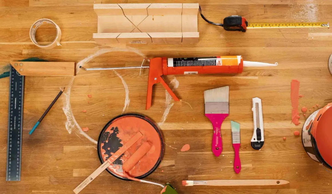 Un tableau peinture acrylique sur bois, prêt pour un projet de bricolage. Pinceaux, rouleau, pot de peinture orange, pistolet à mastic, ruban adhésif, mètre et équerre sont disposés sur un plan de travail en bois.