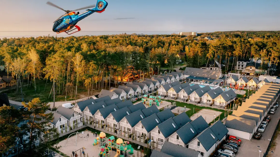 Widok z lotu ptaka na holiday park z domkami, placem zabaw i basenem, a nad nim latający helikopter.