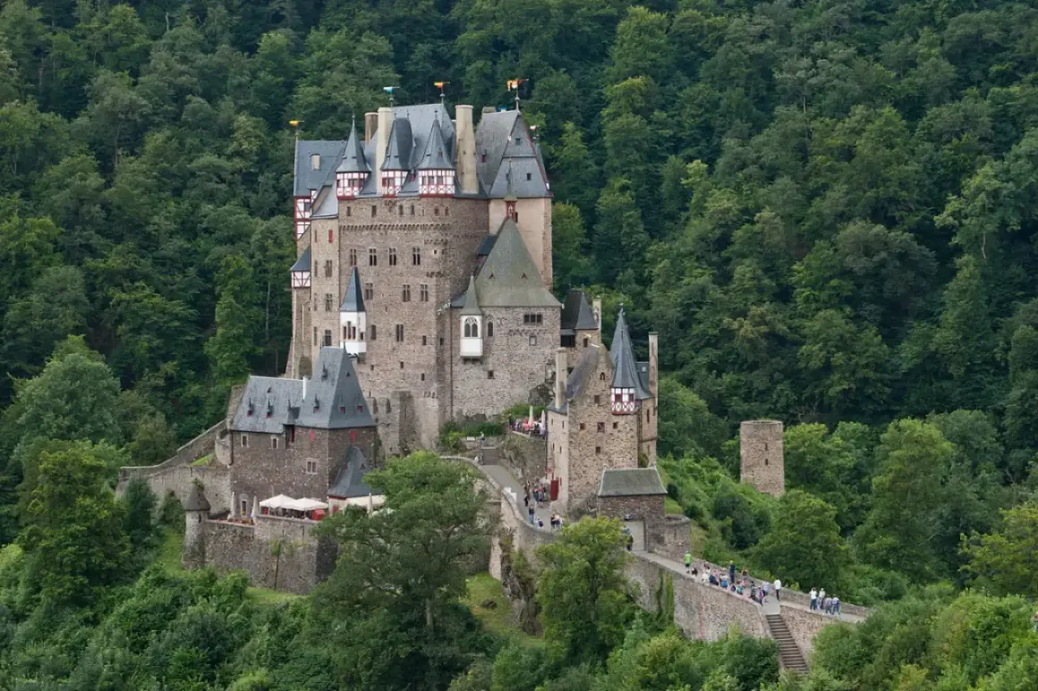 Burg Eltz, ett sagoslott omgivet av grönskande skog, reser sig stolt på en klippa. Många besökare vandrar längs murarna.