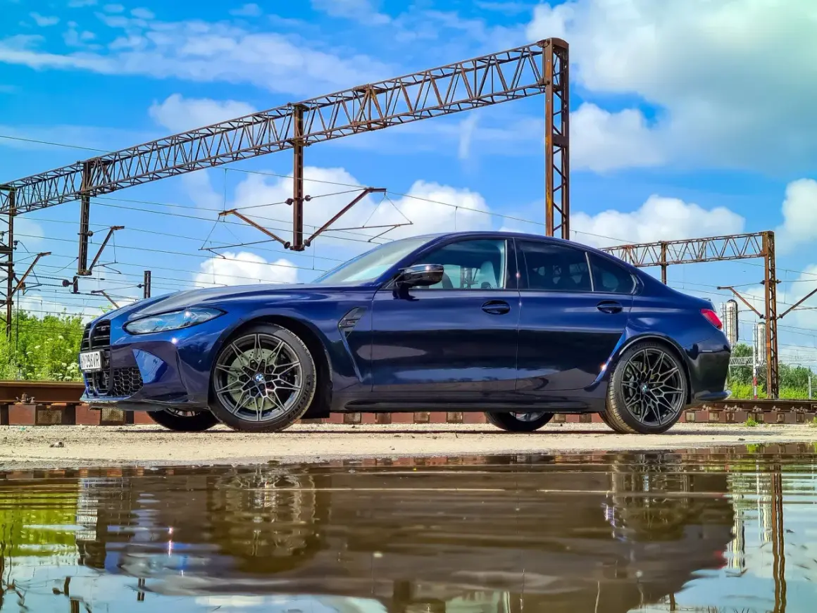 Granatowe BMW M3 G80 stoi obok torów kolejowych, odbijając się w kałuży.
