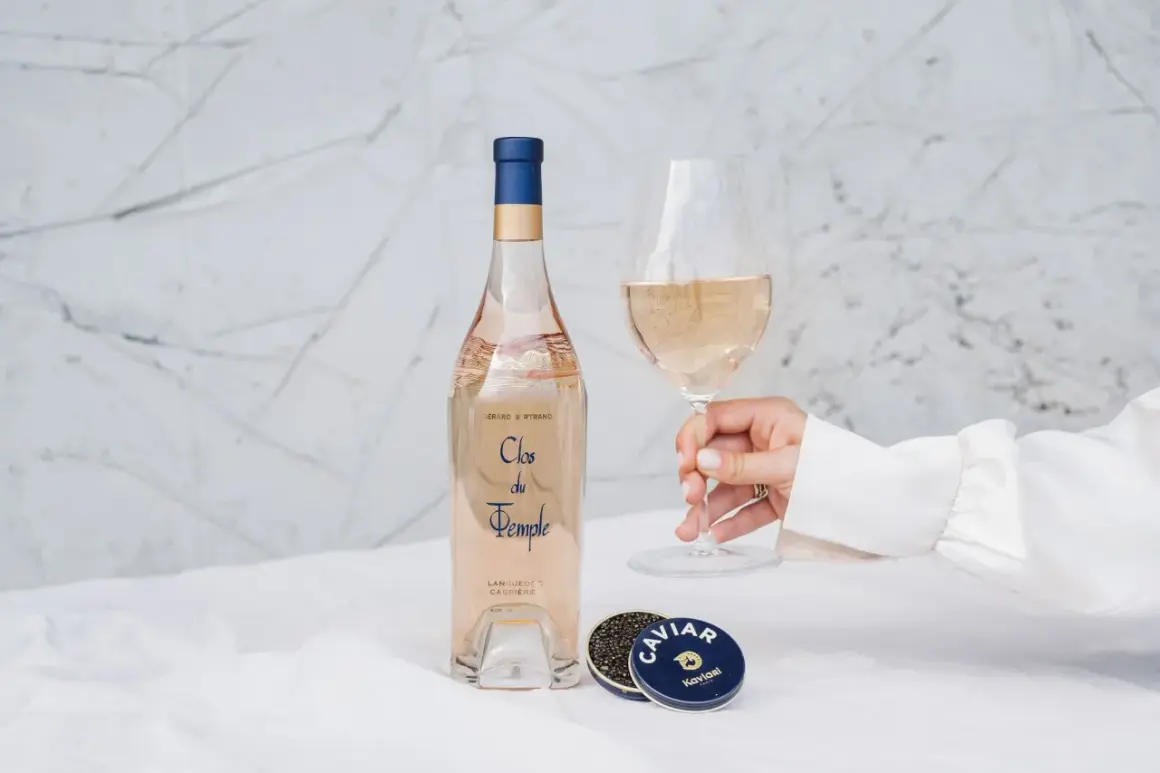 Une main tient un verre de vin rosé, à côté d'une bouteille et de caviar. Le danger du vin rosé est qu'il est si délicieux qu'on en redemande.
