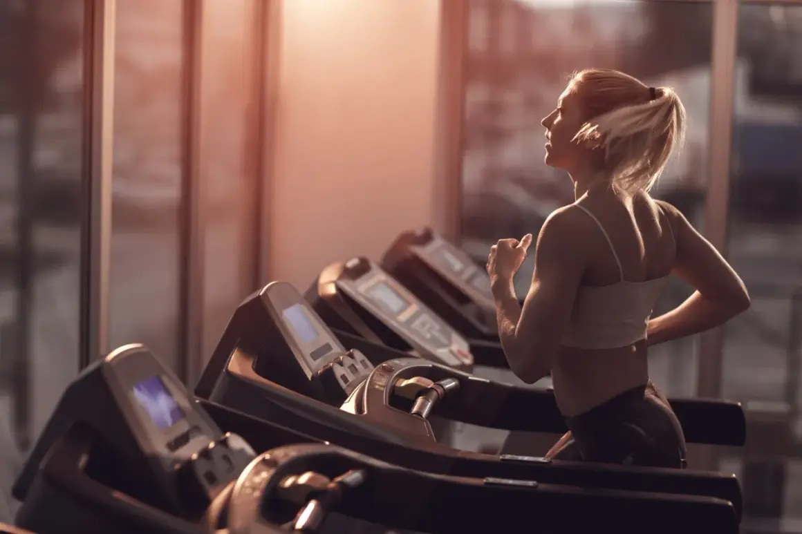 Frau mit blondem Pferdeschwanz walkt auf dem Laufband im Fitnessstudio, das warme Abendlicht fällt durch die Fenster.