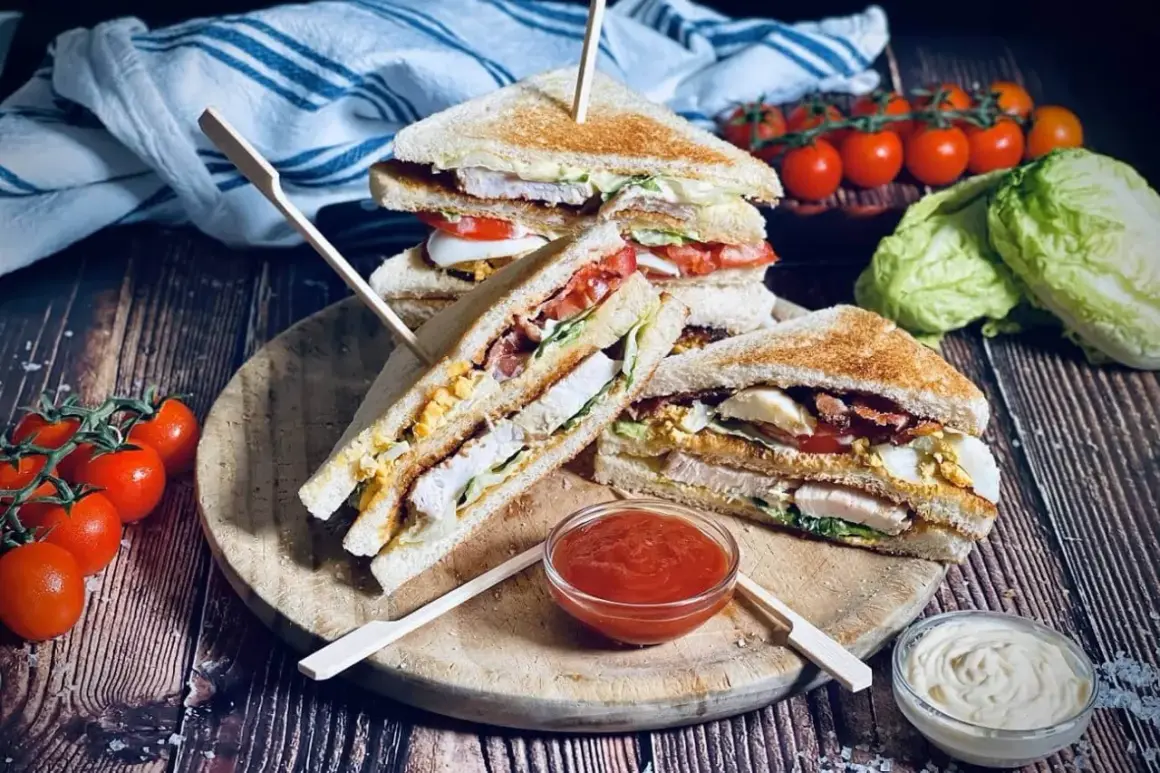 Leckere Sandwich-Ideen: Gestapelte Club-Sandwiches mit Hähnchen, Speck, Ei und Tomaten auf einem Holzbrett, daneben Tomaten und Salat.