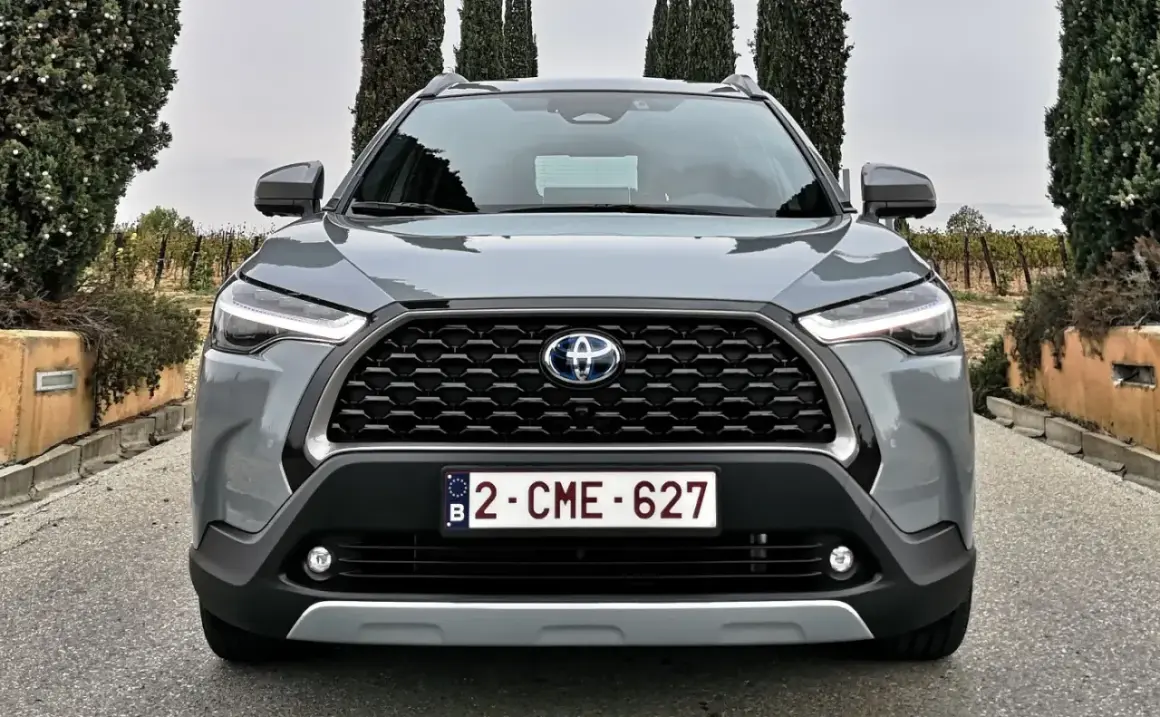 Szary SUV Toyota Hybryda 2022 z przodu, z grillem i światłami LED, na tle winnicy i drzew.