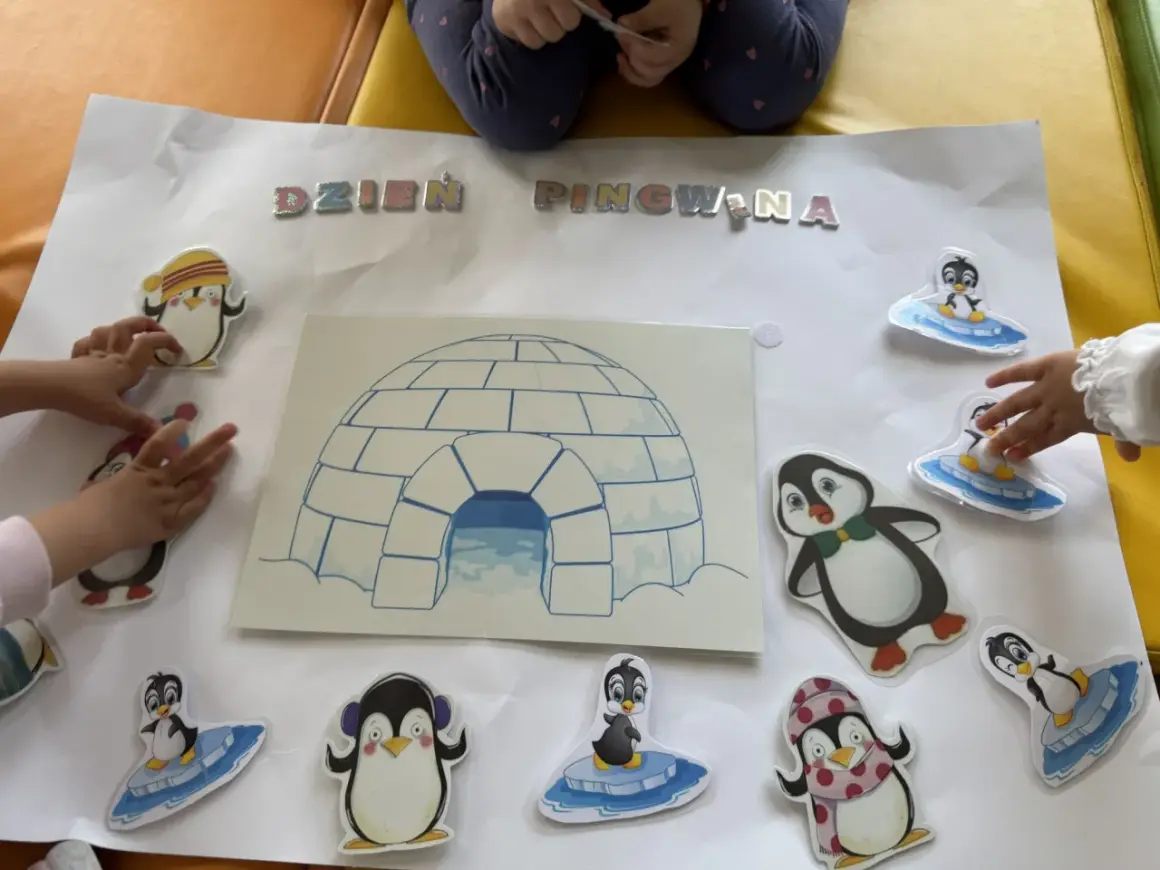 Dzieci tworzą plakat na dzień pingwina w przedszkolu, ozdabiając go naklejkami pingwinów i igloo.