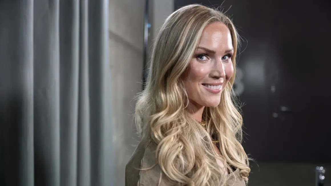Marie Serneholt ler mot kameran med sitt blonda hår fallande i lockar. Hon bär en beige jacka och ett guldigt halsband.