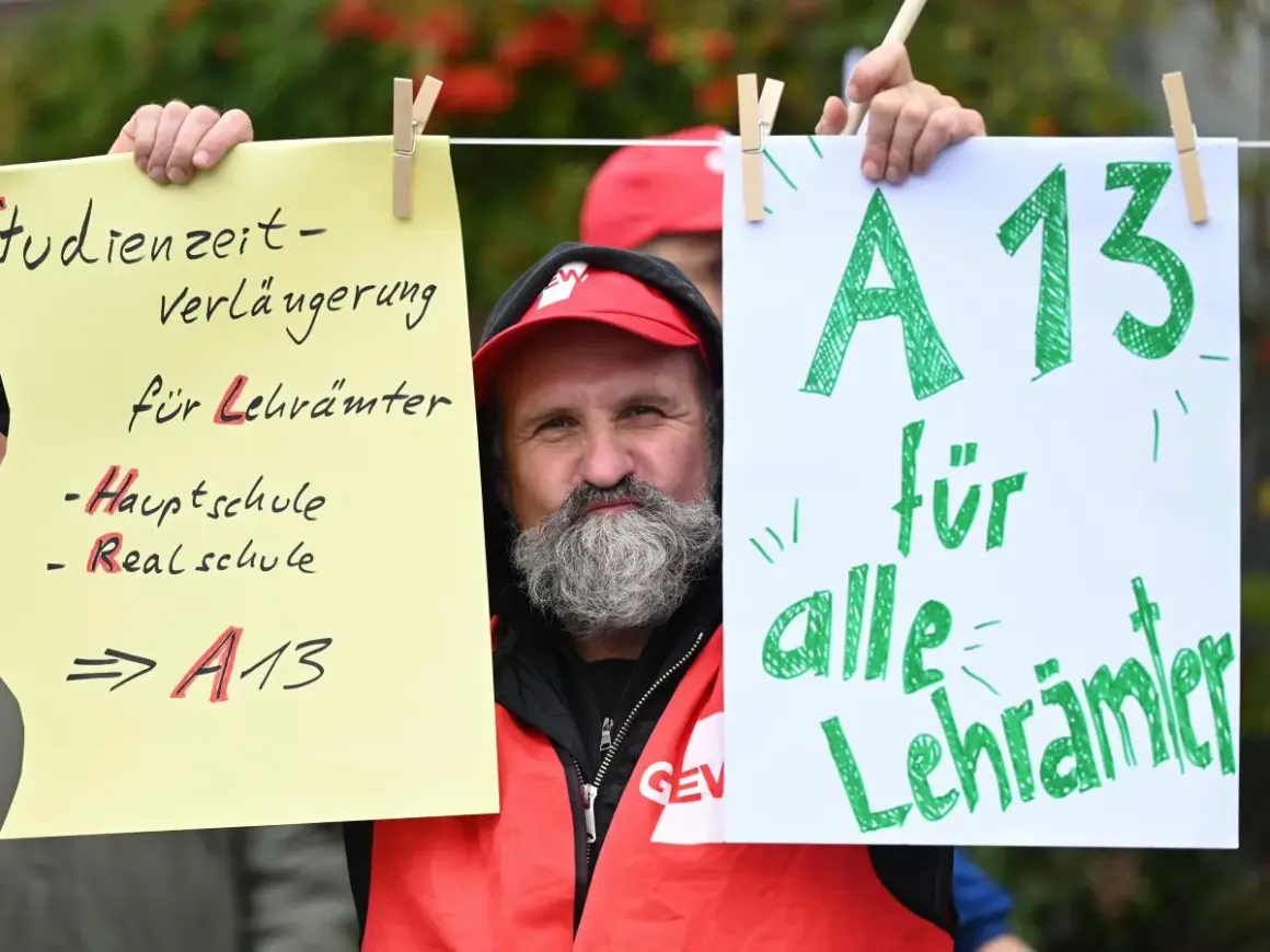 Mann mit Bart und roter Weste hält Schilder hoch. Ein Schild fordert "A13 für alle Lehrer" und thematisiert das Lehrer Gehalt Berlin.