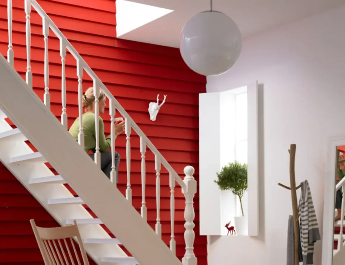 Ein rotes, holzvertäfeltes Treppenhaus mit weißer Balustrade. Eine Frau sitzt auf der Treppe. Ein Hirschkopf und ein Baum schmücken die Wand. So lässt sich ein Treppenhaus gestalten.