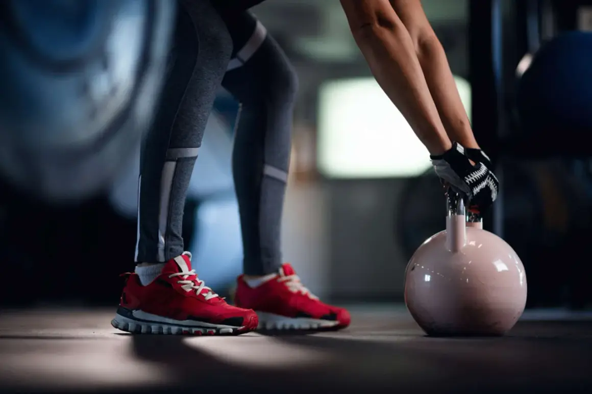 Person mit roten Schuhen und Trainingshandschuhen hebt abends eine Kettlebell für das Krafttraining.