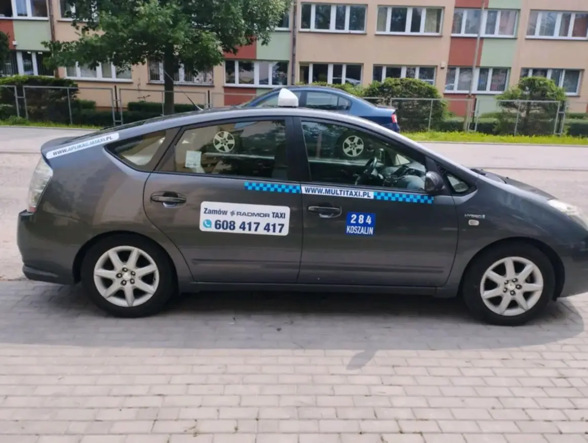 Szary hybrydowy Prius z napisem "284 KOSZALIN" i numerem telefonu do zamówienia taksówki.