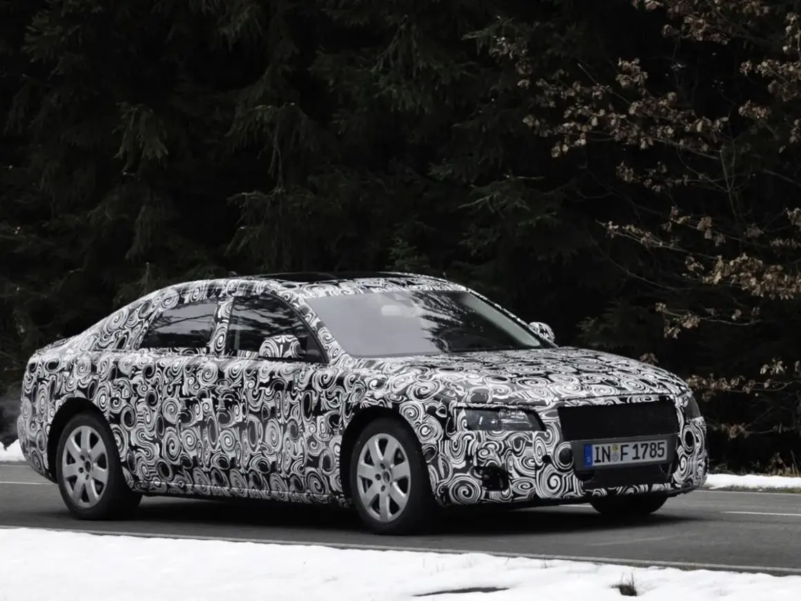 Audi Limousine mit Tarnfolie fährt auf schneebedeckter Straße vor dunklem Wald.