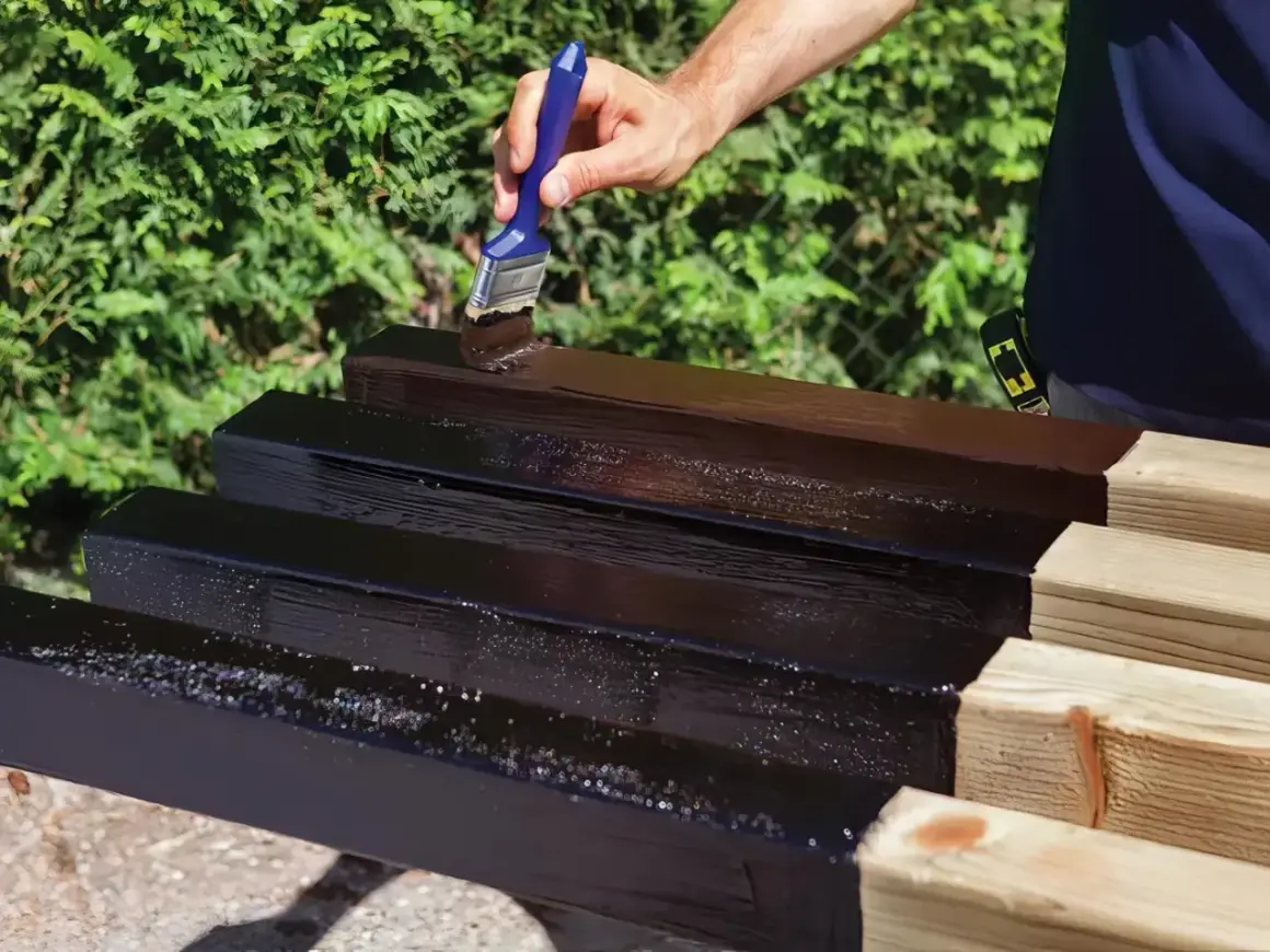 Mano che applica vernice scura su listelli di legno grezzo, un passo essenziale per imparare come impermeabilizzare il legno e proteggerlo.