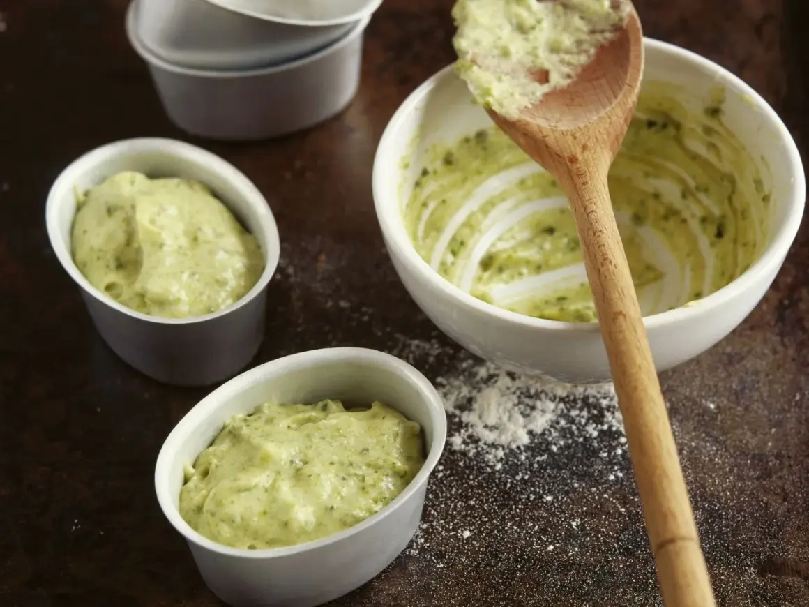 Kleine Förmchen werden mit einer grünen Masse gefüllt, die nach Pistaziencreme selber machen aussieht. Ein Holzlöffel liegt in einer Schüssel mit der Masse.