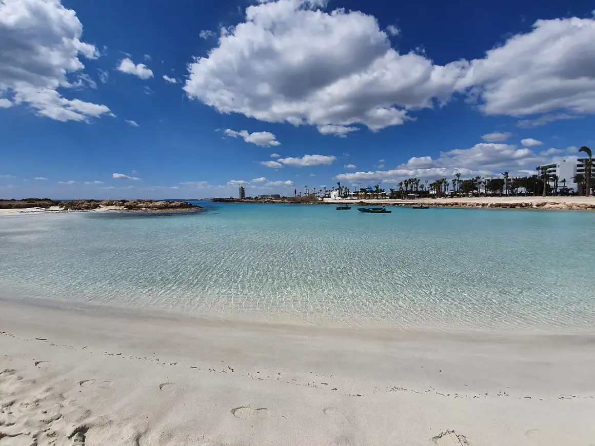 Kristallklart vatten och vit sand vid Nissi Beach, Cypern. Små båtar guppar i det turkosa vattnet under en blå himmel med vita moln.