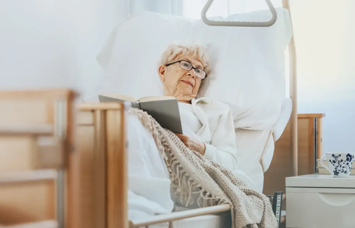 Ältere Dame liest im Bett, eine Auszeit in der Kurzzeitpflege ohne Pflegegrad. Gemütlich eingekuschelt mit einem Buch.