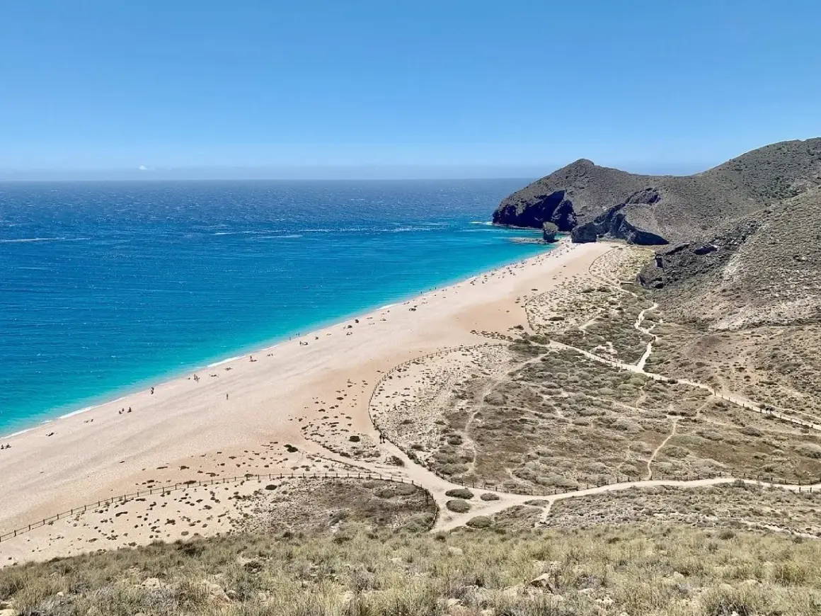 Una cala de arena dorada se extiende junto a un mar turquesa. Senderos serpentean por las colinas áridas que rodean la playa.